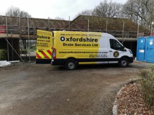 A yellow van for Oxfordshire Drain Services parked near a construction site with scaffolding, offering drain surveys and water jetting services.