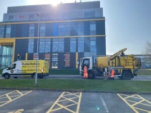 A yellow drain service truck parked outside a modern building, with workers in orange vests inspecting a drainage cover.