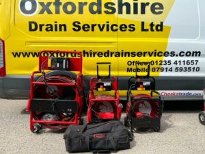 A display of drain cleaning equipment, including three red machines and a black tool bag, against the backdrop of an Oxfordshire Drain Services van.
