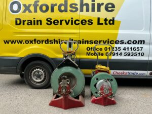 Two drain cleaning machines in front of a yellow van labeled "Oxfordshire Drain Services Ltd" with contact information.